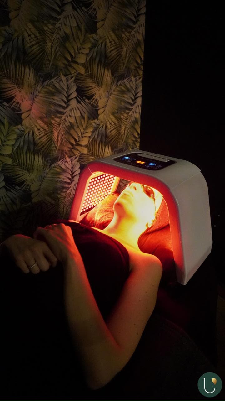 Persona recibiendo tratamiento facial con terapia de luz LED roja bajo un dispositivo de arco blanco.