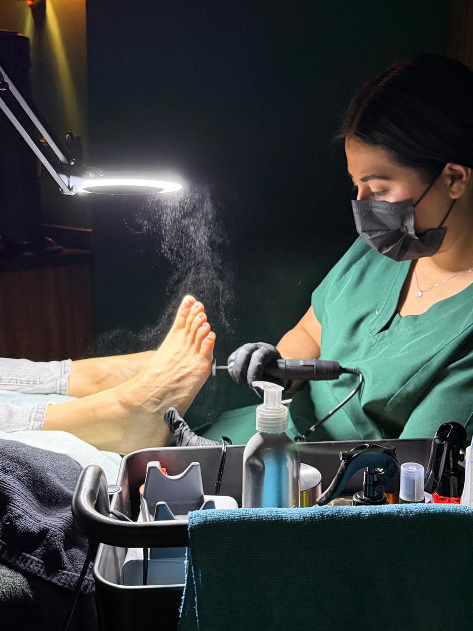 Especialista con mascarilla realizando una pedicura con torno eléctrico bajo una potente luz anular.