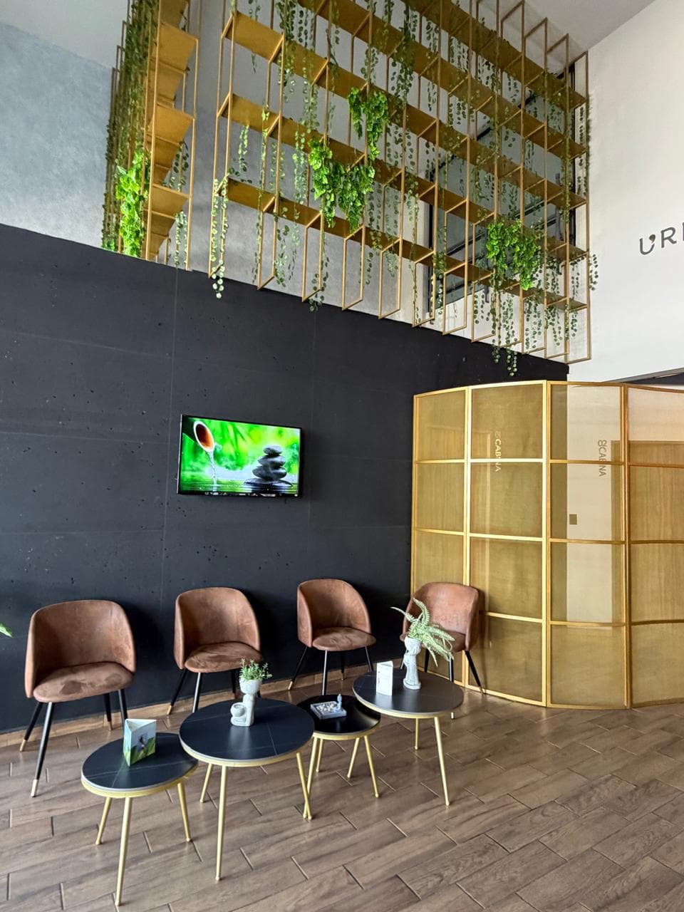 Sala de espera moderna con sillas marrones, mesas negras y plantas colgantes en rejilla dorada.
